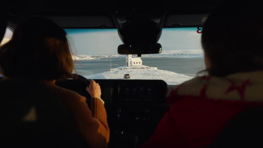 An over-the-shoulder view from inside a car of two people driving towards a remote, solitary church in a vast, snowy landscape