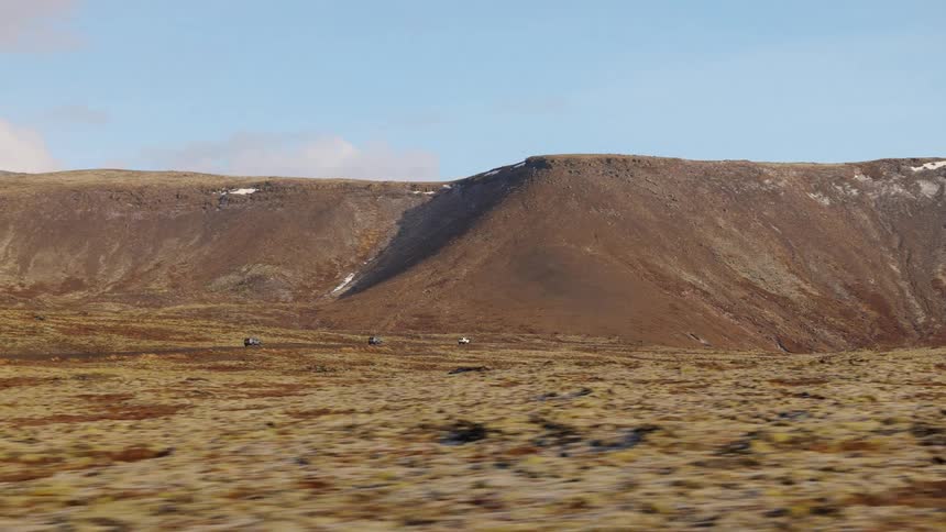 Traveling through a vast, barren tundra landscape with a large, rocky hill and patches of snow under a clear sky