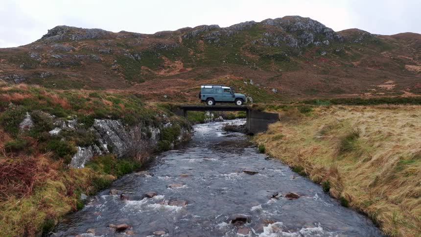 An off-road vehicle crosses a remote bridge over a river flowing through a rugged mountain valley