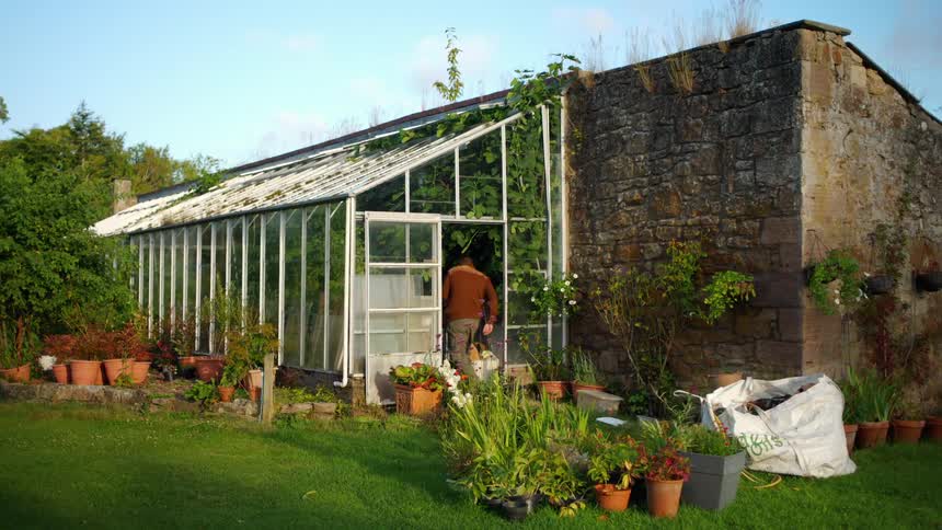 A male gardener carries a basket as he walks into a rustic greenhouse next to a stone wall