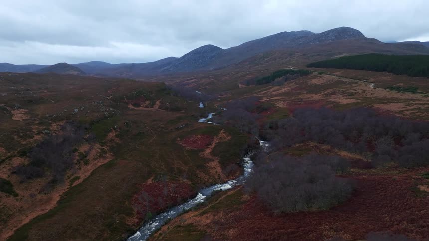 An aerial view captures a river flowing through a vast, rugged mountain valley on an overcast day