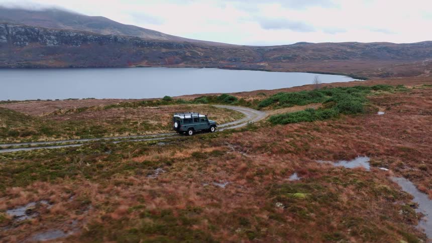 An aerial view orbits a Land Rover Defender parked on a rugged dirt track beside a tranquil loch in the remote Scottish Highlands