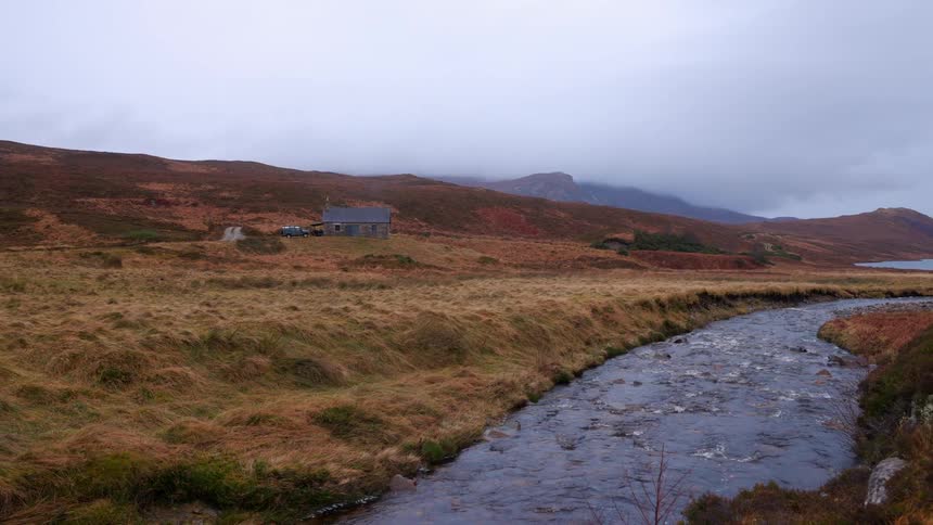 A lone stone cottage sits by a flowing river in the vast, remote landscape of the Scottish Highlands on an overcast day