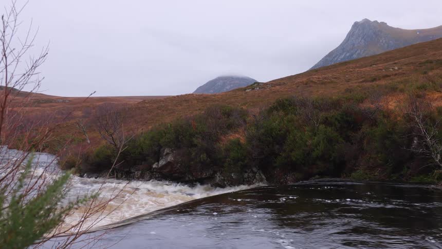 A powerful river cascades over a ledge into churning rapids in the vast, moody landscape of the Scottish Highlands on a cloudy day