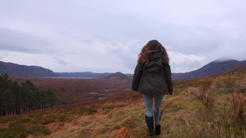 A woman with long hair blowing in the wind walks in slow motion through a vast, grassy highland valley surrounded by mountains