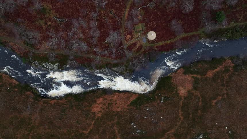 An aerial top-down view of a river with strong rapids flowing through a rugged, autumnal landscape with a lone yurt on its bank