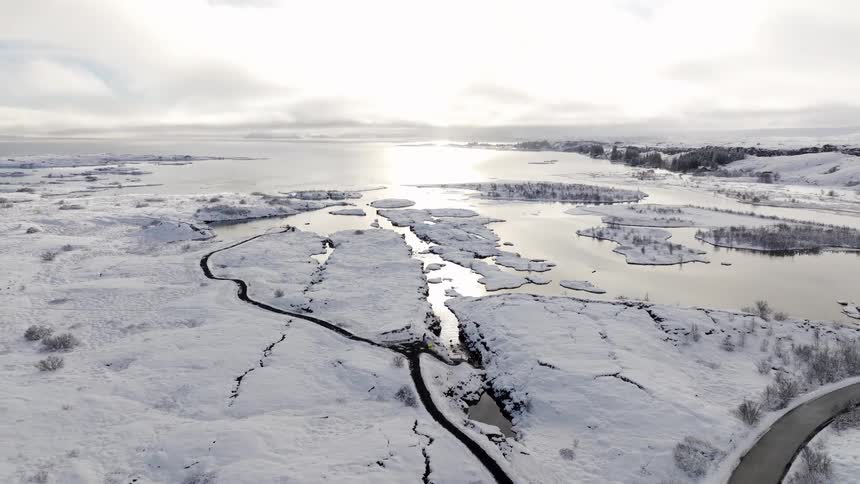 A stunning high-angle aerial view of a vast, snowy Icelandic landscape with the sun reflecting off a partially frozen lake