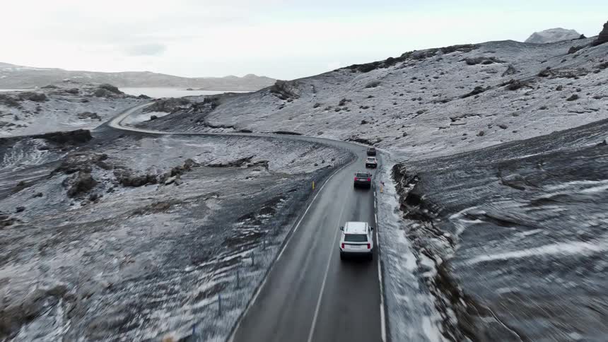 An epic aerial view follows a convoy of cars navigating a winding road through a desolate, snow-covered volcanic landscape