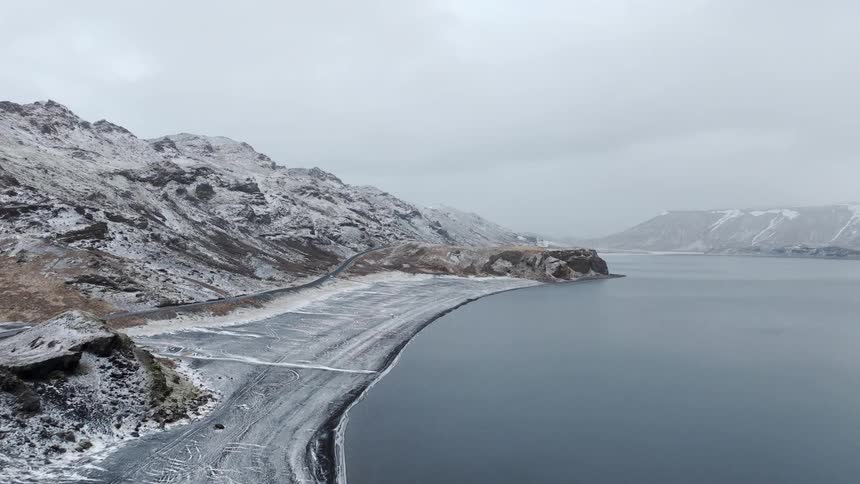 A serene aerial shot captures a snow-covered black sand beach along a tranquil lake surrounded by mountains on a cold, overcast day