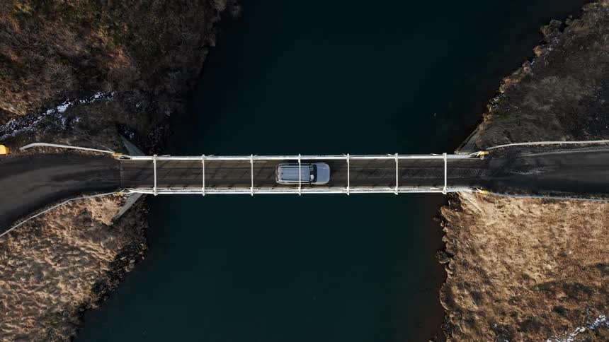 A stationary aerial top-down view of a silver SUV parked in the middle of a bridge over a dark river in a rugged landscape