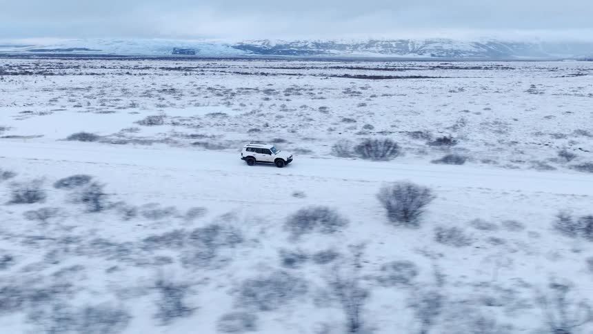 A drone follows a white SUV as it drives down a remote road through a vast, snow-covered landscape