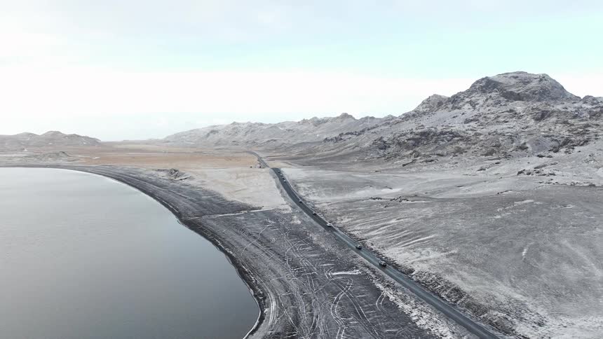 High aerial view of cars traveling on a scenic coastal road through a vast, snow-dusted mountainous landscape in winter