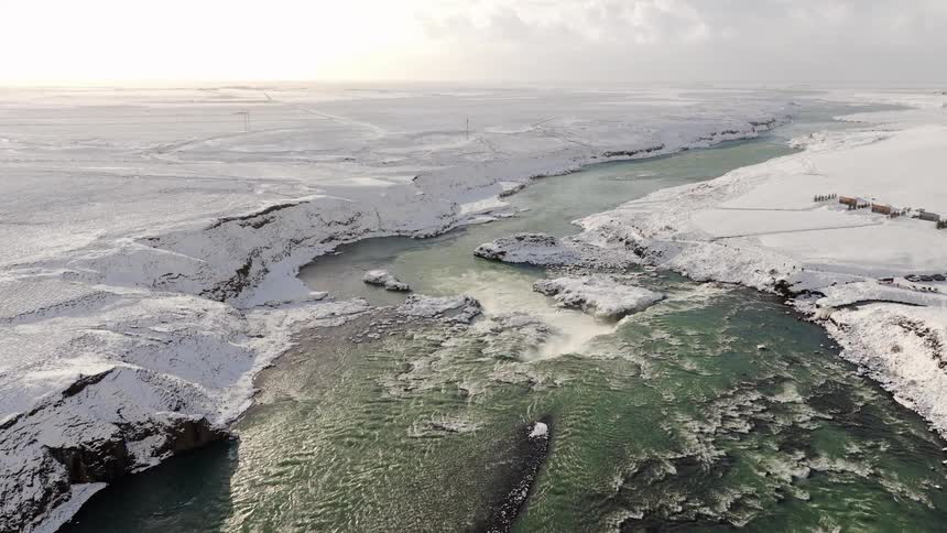 A stunning aerial view shows a powerful waterfall cascading through a vast, snow-covered landscape in Iceland during winter