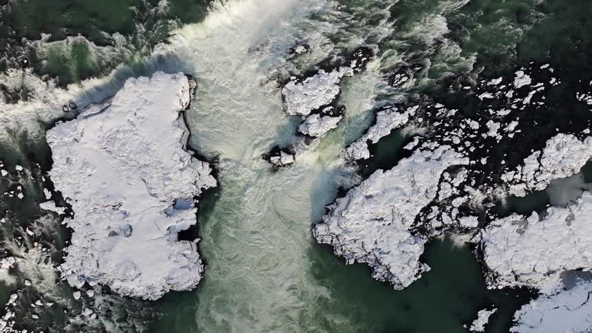 A rising top-down aerial view of a powerful winter waterfall churning through a snowy, rocky canyon