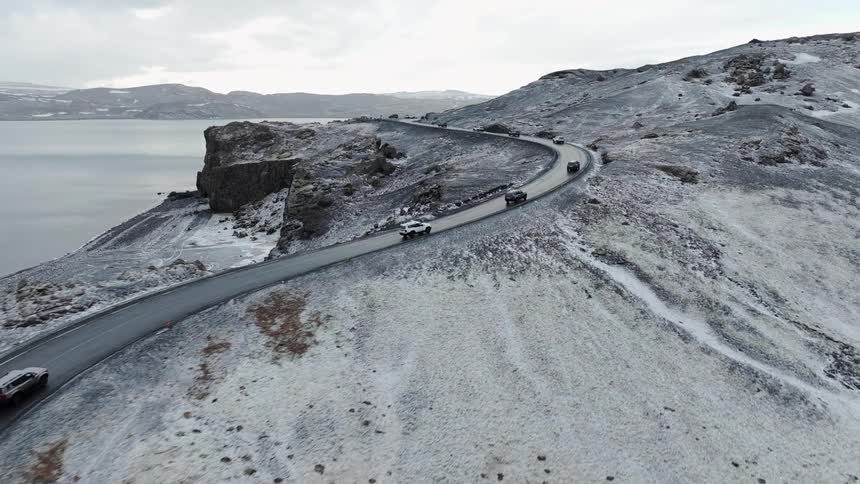 An aerial view captures a convoy of cars driving along a winding coastal road through a vast and snowy Icelandic landscape