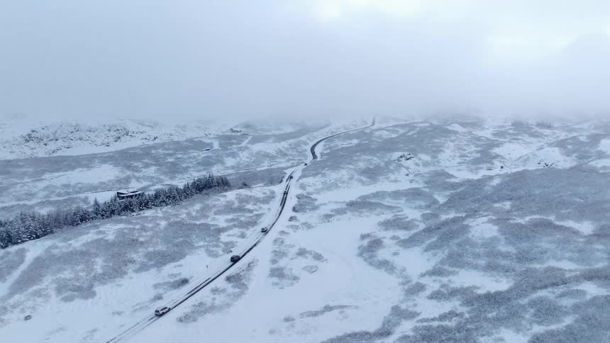 A drone slowly ascends over a snow-covered mountain landscape, revealing cars driving along a winding road in foggy conditions