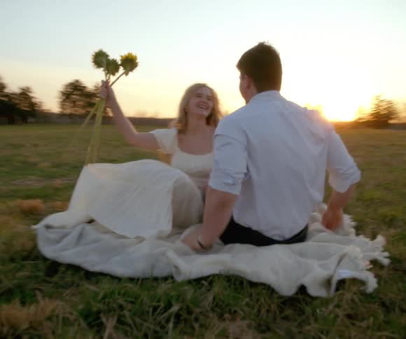 A happy young couple laughs together on a blanket in a grassy field at sunset as the woman playfully holds sunflowers