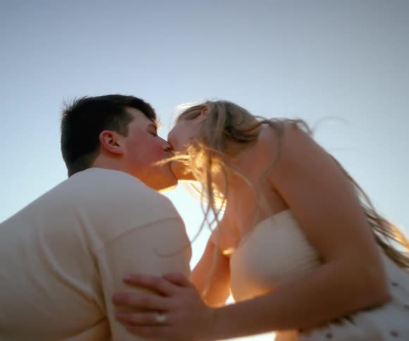 A loving couple shares a romantic kiss while leaning on a fence in the country, backlit by a beautiful golden sunset