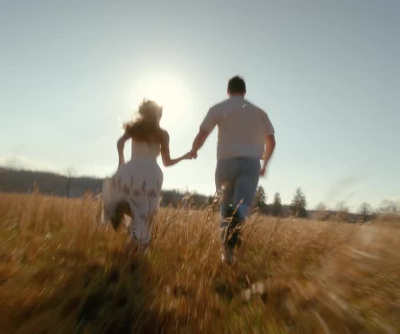 A young romantic couple holds hands and runs through a golden grassy field towards the bright sun