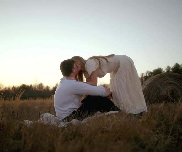 An affectionate young couple shares a romantic kiss in a grassy field at sunset, with a hay bale in the background