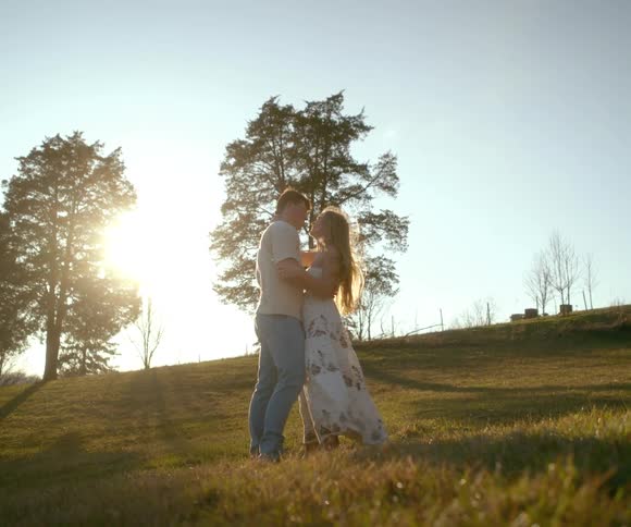 A romantic young couple embraces and kisses on a windy hillside as the sun sets behind them