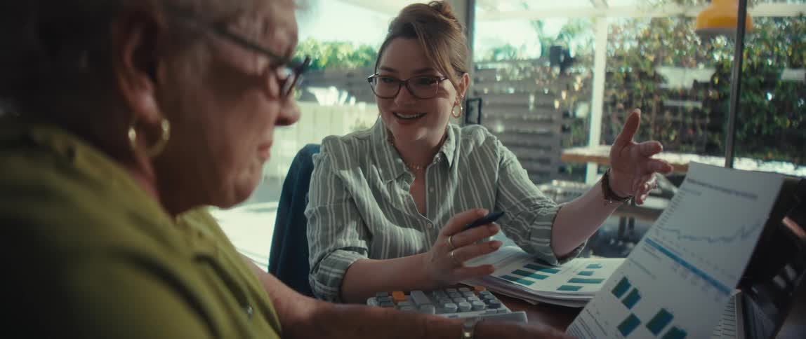 A young female financial consultant enthusiastically explains investment charts to a senior woman during a home meeting