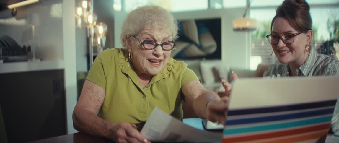 A cheerful elderly woman laughs and points at a laptop screen while a younger woman helps her at the dining room table