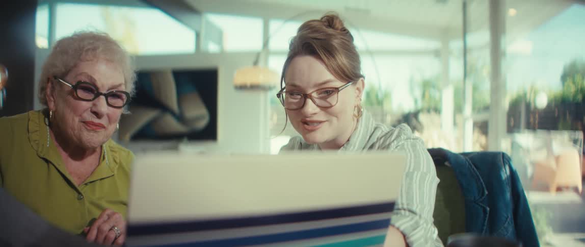 An adult daughter joyfully assists her elderly mother with a task on a laptop, and they share a happy laugh in their sunlit home