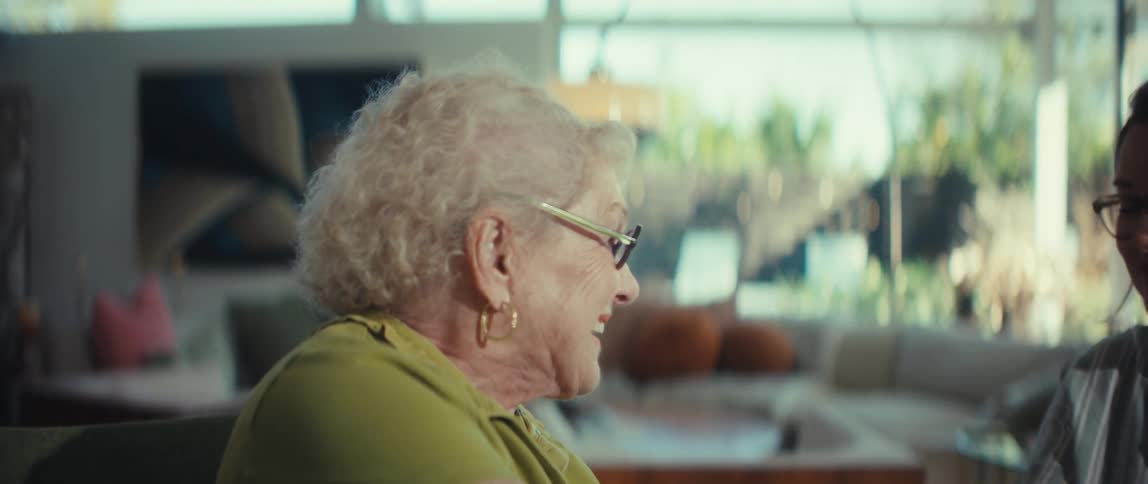 A happy senior woman wearing a hearing aid holds hands and laughs with her younger companion in a sunlit living room