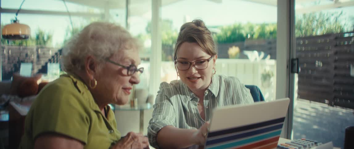 A caring young woman helps a senior woman use a laptop, sharing a happy and surprising moment together in a modern, sunlit home