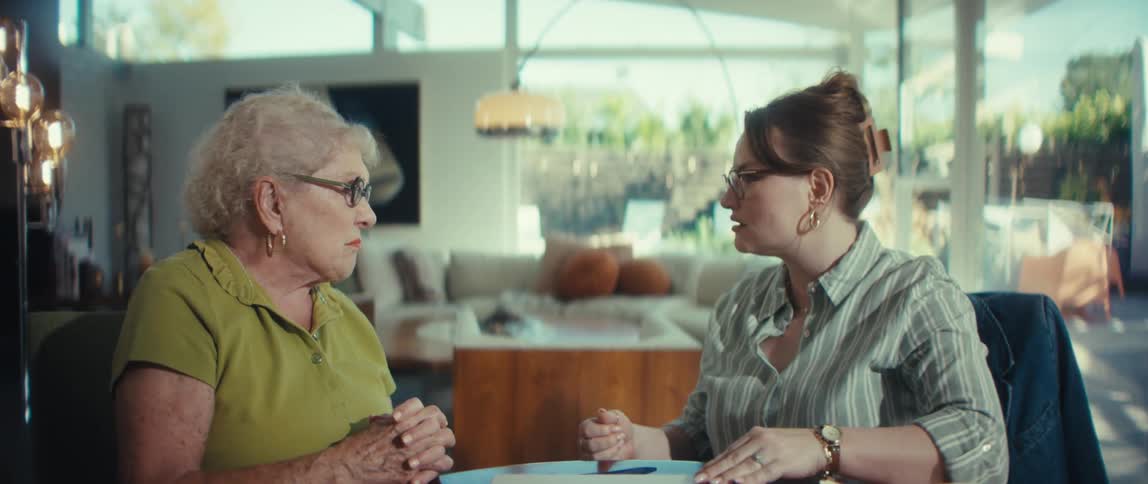 A young professional woman explains financial options on a laptop to a senior woman in her modern home, ending with a firm handshake