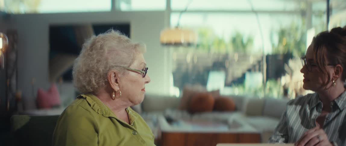 A young woman and an elderly woman hold hands and share a warm, smiling conversation in a sunlit living room
