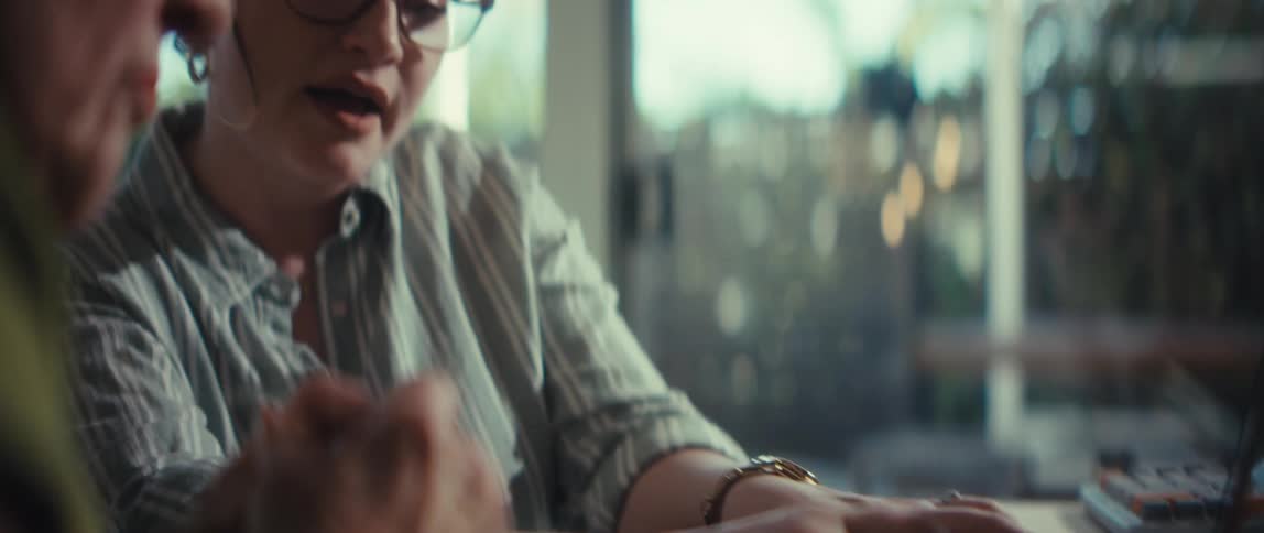 A young woman wearing glasses smiles and talks to a person sitting next to her while working on a laptop