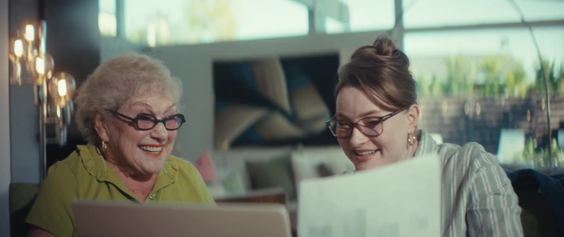 A cheerful elderly woman and her helpful granddaughter laugh together while looking over documents and a laptop in a stylish, sunlit room
