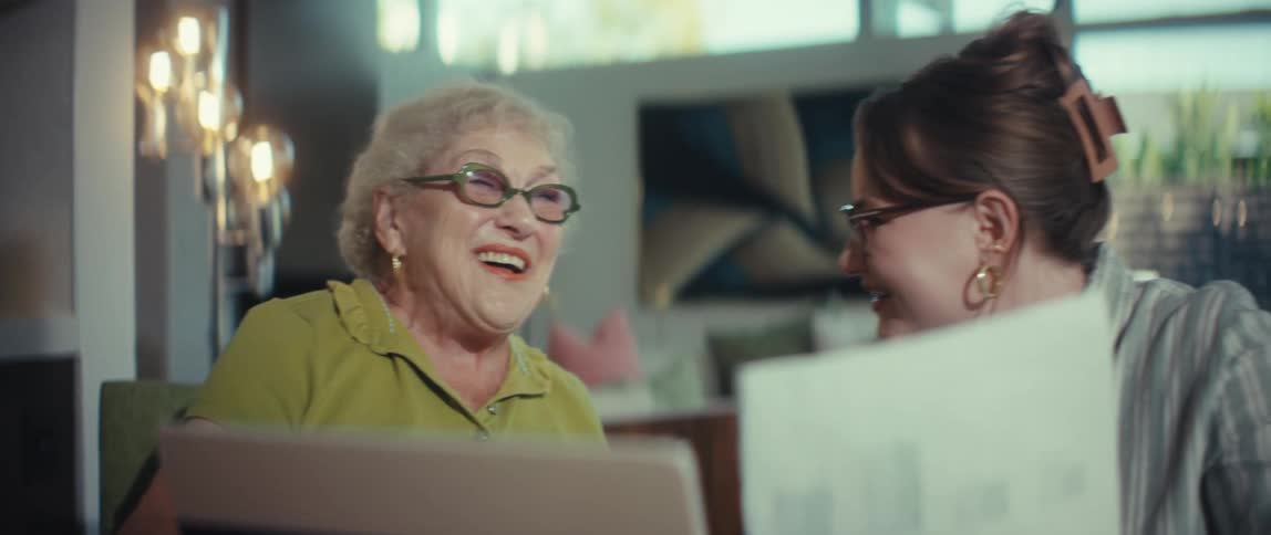 A cheerful senior woman and her younger companion share a laugh while reviewing financial documents together at a table