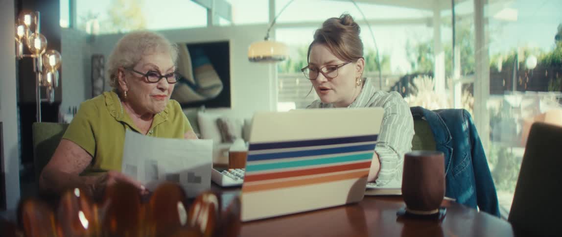 A helpful young woman assists an elderly woman with financial planning, reviewing documents and using a laptop at their sunny dining room table