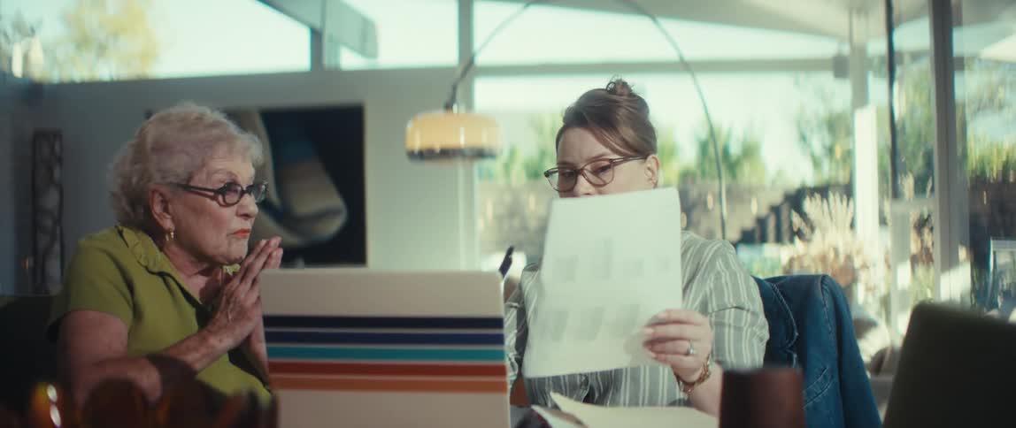 A young professional patiently explains financial documents to a concerned senior woman in a bright, mid-century modern home