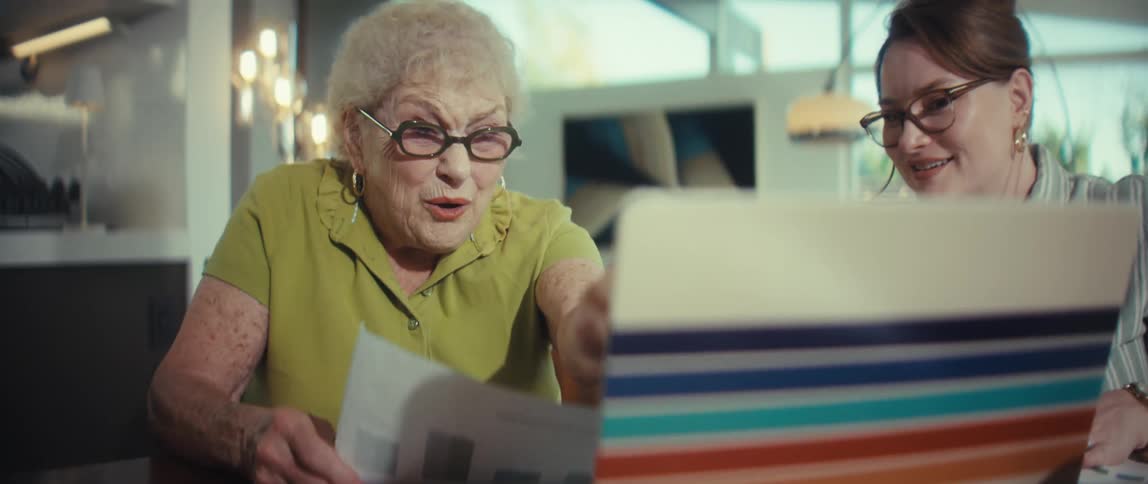 A happy elderly woman and a younger woman review a successful report with a chart, laughing and pointing at a laptop screen in a modern home