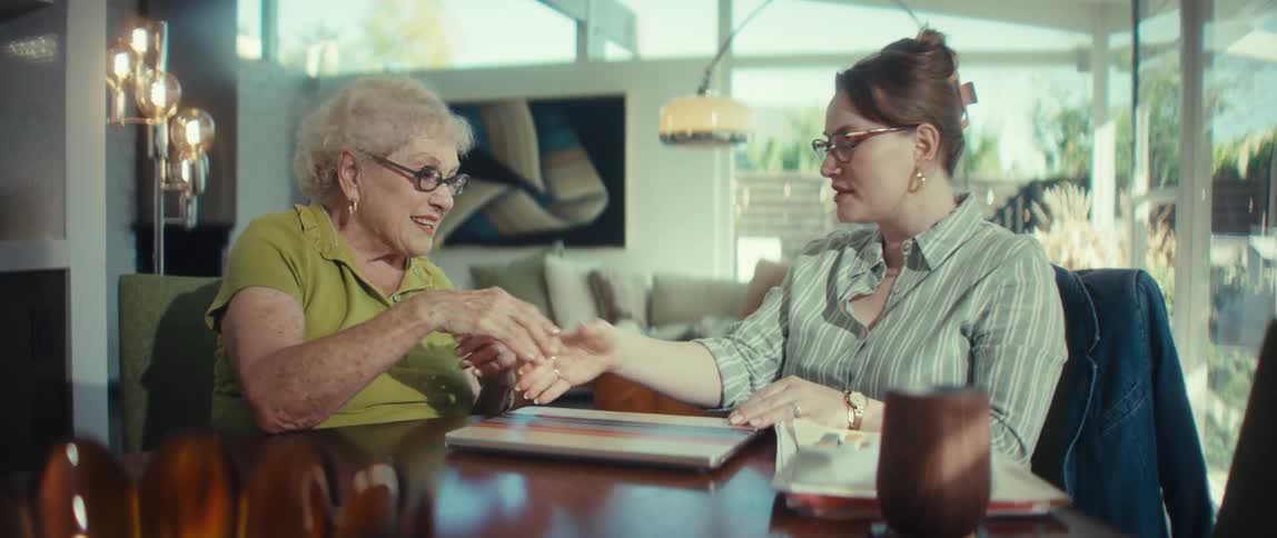 A younger woman shakes hands with a smiling senior woman before they look at a laptop together at a dining table in a modern home