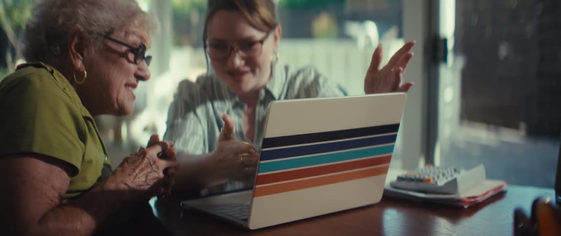 A younger woman opens a laptop and explains something to an elderly woman as they sit together at a table in a sunlit room