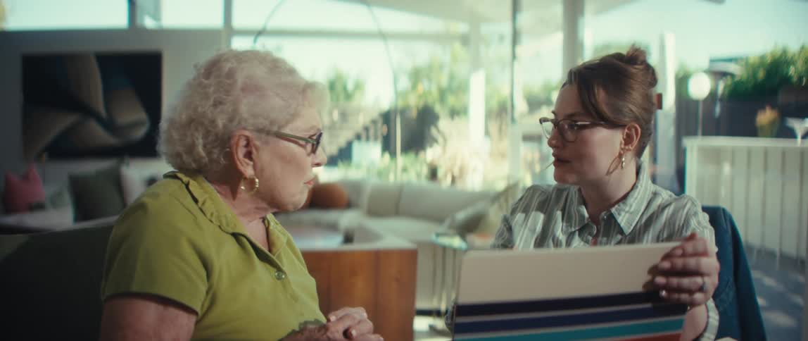 A female advisor compassionately explains documents to a senior woman in her modern home, holding her hand for reassurance