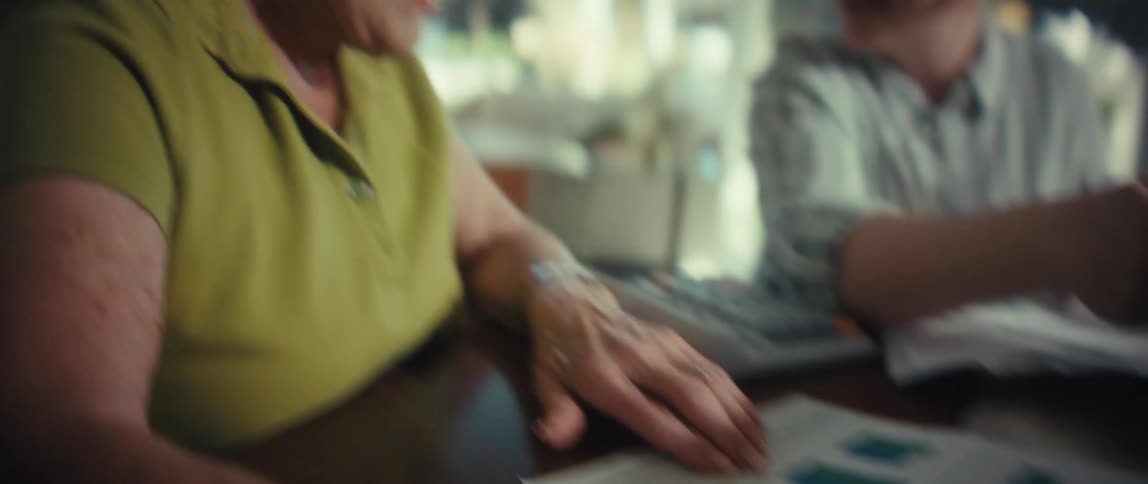 A caring young woman with glasses helps a smiling senior woman with her finances, reviewing documents together at a table in a sunlit room