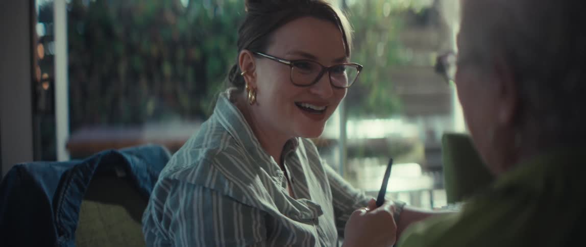 A smiling woman with glasses has a joyful and animated conversation with an elderly person in a naturally lit room