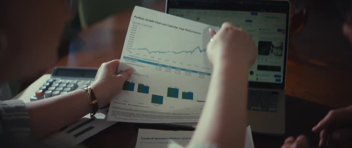 Over the shoulder view of two people analyzing stock market data and portfolio growth on a laptop and printed charts