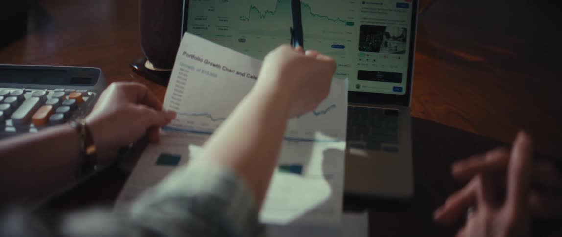 An over-the-shoulder shot of two people comparing a portfolio growth chart on paper to a live stock market chart on a laptop