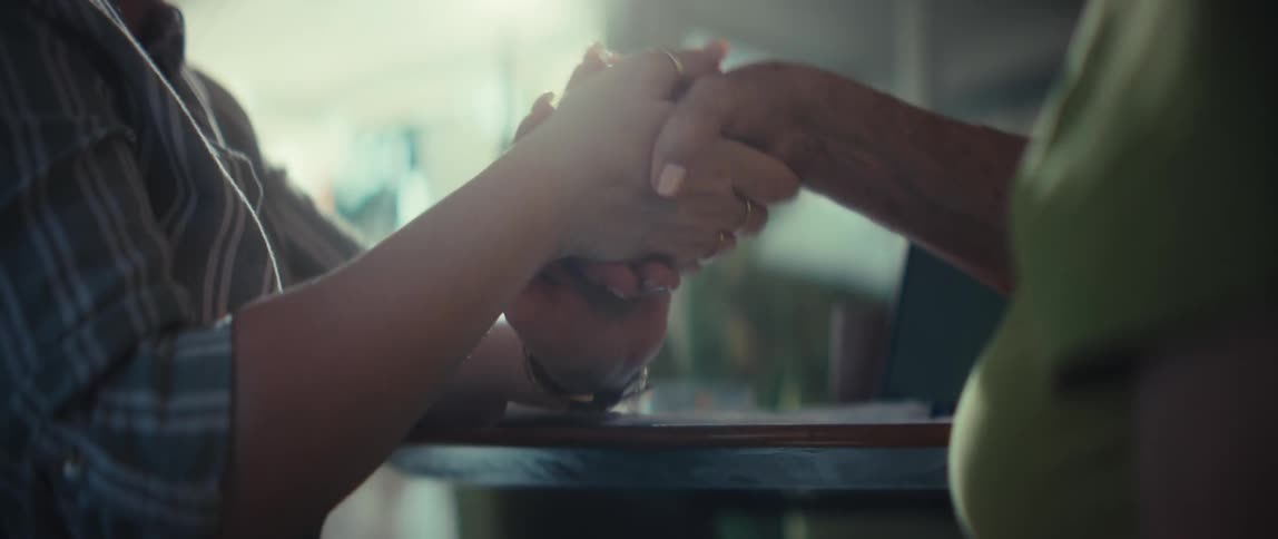 A young woman offers comfort and support to an elderly woman by tenderly holding her hands across a table in a sunlit room