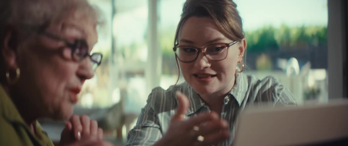 A young woman with glasses enthusiastically explains something on a laptop to a senior woman as they sit together at a dining table in a sunlit room