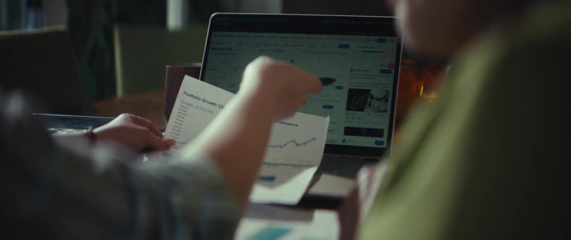 Over-the-shoulder shot of two people analyzing their investment portfolio performance using a laptop and printed charts
