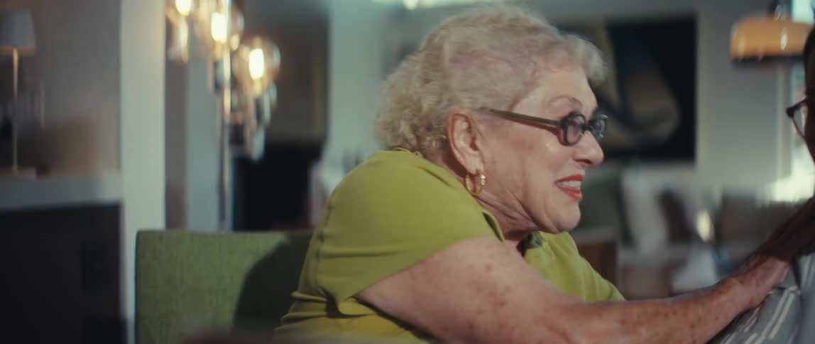 An expressive elderly woman claps and laughs with joy as a younger woman shows her some documents in a modern room