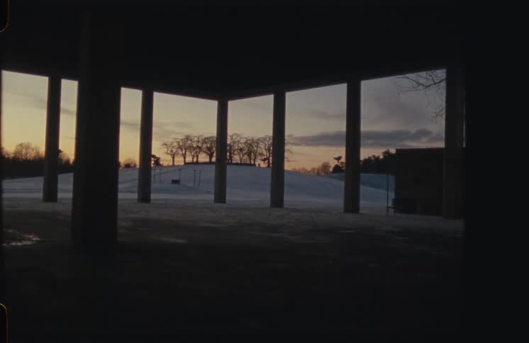 A still shot from under a concrete structure frames a snowy hill with silhouetted trees against a sunset sky